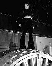 Chloe Chaidez standing on top of a grey vault door, wearing a black crop top and black trousers