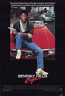 Axel Foley (portrayed by Eddie Murphy) sits on the hood of a red Mercedes-Benz convertible with a pistol pointing out on his left hand and his left foot resting above the word "Hills". The caption above reads "He's been chased, thrown through a window, and arrested. Eddie Murphy is a Detroit cop on vacation in Beverly Hills."