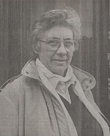Black and white photograph of the head and shoulders of a woman with short hair and large glasses wearing a trench coat over a dark v-necked sweater and a white cowl-necked blouse.