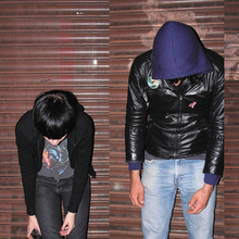 The album cover combines two images of Crystal Castles members — one of Alice Glass and another of Ethan Kath — standing in front of a metallic shutter. On the left, Glass has black hair, a black jacket, and jeans, looking down with her arms hanging by her sides. On the right, Kath is wearing a hooded jacket with the hood up and blue jeans, also looking down with his arms hanging by his sides.