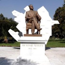 Picture of the statue of Aliyev, a bronze sculpture placed on a marble pedestal. Aliyev wears a suit and is sat on a chair with his right leg crossed. The pedestal has written "Heydar Aliyev 1923 - 2003" in capital golden letters. Behind the statue, there is a sculpture of Azerbaijan, which features the Spanish names of some Azerbaijan cities.