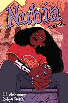 Nubia sits outside a white house wearing a Wonder Woman logo t-shirt and has long natural hair. She wears gray pants and has polka dot fingernail polish.
