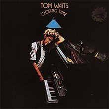 A man leans against a piano in a dark room. The arched text above him reads "Tom Waits Closing Time".