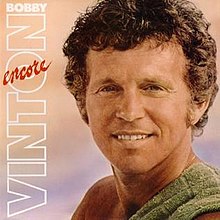 A photo of Bobby Vinton from the shoulders up smiling to his left at the camera. He is bare-chested with a green towel over his left shoulder. Beside him are the words BOBBY VINTON in white and ENCORE in red. The background is peach on the top, blue on the bottom, like a beach sunset.