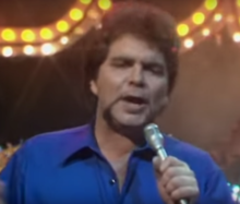 A 34-year-old man is shown in an upper body shot. He is singing into a microphone held in his left hand. He wears an open blue shirt. His hair is in an "afro" cut, while his beard shows cleared upper lip and lower lip with a line of beard framing his mouth and lower chin. Behind him are yellow and white lights (partly obscured).