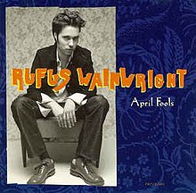 A black-and-white photograph of Rufus Wainwright appears atop a square, dark-blue paisley-patterned background; atop both items is the singer's name in an all-caps orange font, above "April Fools" in a much smaller white font.