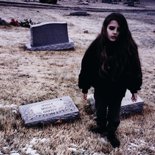 A young person with long dark hair stands in a cemetery. They are dressed in dark clothing and looks directly at the camera with a serious expression. Two gravestones are visible in the image, one standing upright and another lying flat on the ground. The ground is covered with light frost or snow.