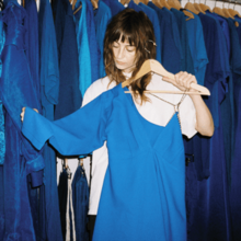 A "girl next door" with long brown hair and a white t-shirt takes a blue dress off its hanger. A rack of similar dresses forms the backdrop.